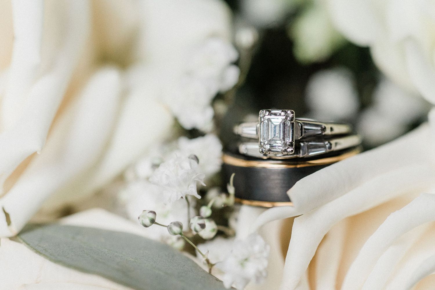 Elegant emerald cut diamond engagement ring with baguette side stones displayed on wedding bands among white flowers.