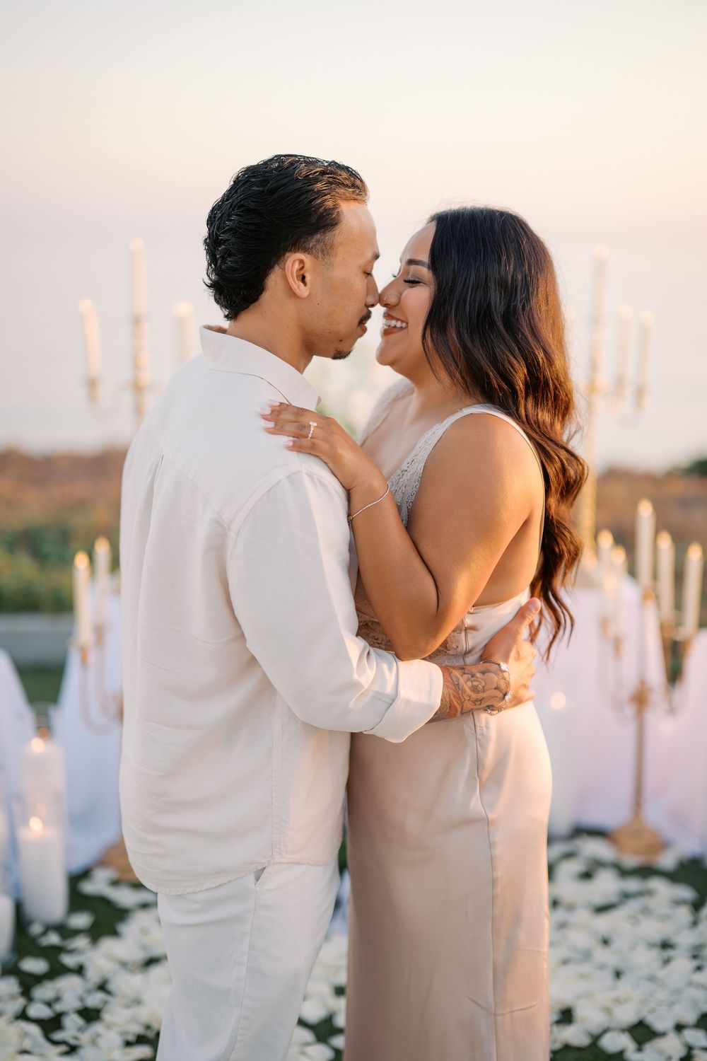 Couple embraces and nearly kisses surrounded by candles and white floral arrangements at a romantic oceanside proposal.