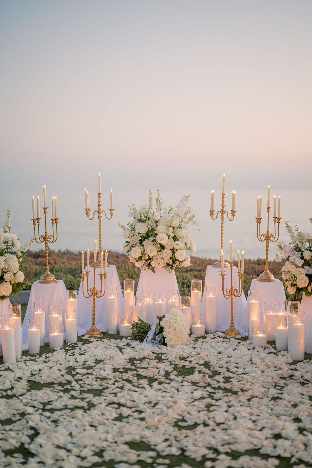 Elegant outdoor wedding altar with gold candelabras, white pillar candles, floral arrangements, and rose petals at sunset.