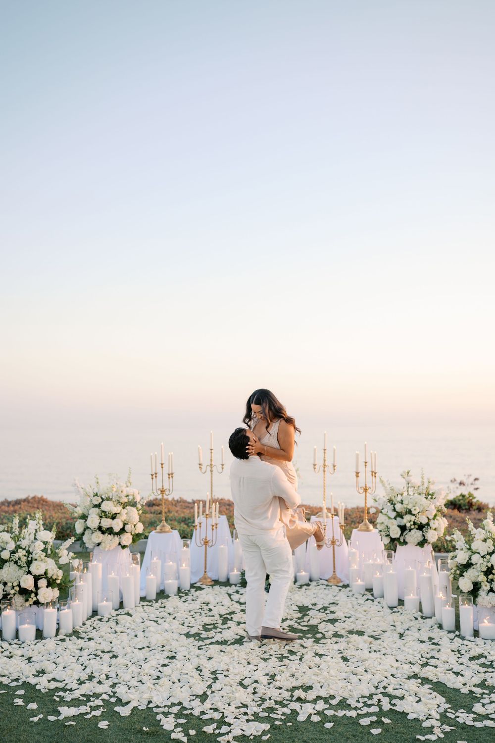Man lifts woman in white amid candles and flowers at sunset proposal.
