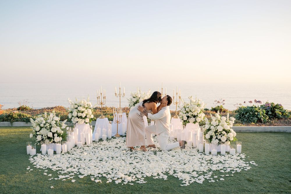 Couple sharing romantic kiss surrounded by white floral arrangements and rose petals at outdoor wedding ceremony at sunset.