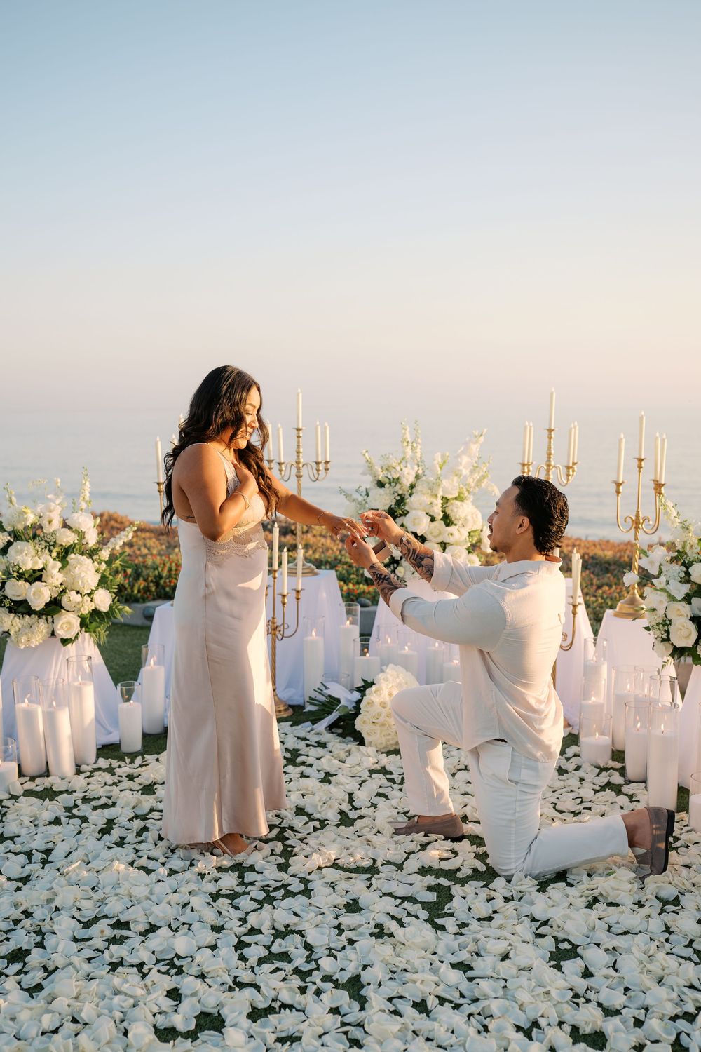 Romantic seaside proposal with man on one knee offering ring to woman surrounded by white petals and candles at dusk.