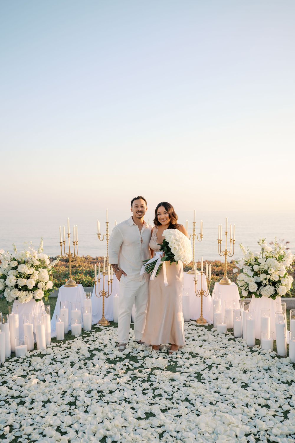 Couple stands smiling among white floral arrangements, candles, and gold candelabras at an outdoor proposal setup.