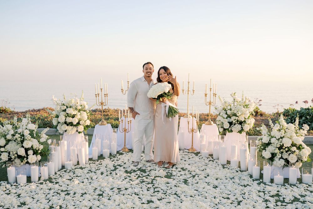 Couple poses at elegant outdoor wedding ceremony with white floral arrangements, candles, and rose petals at sunset.