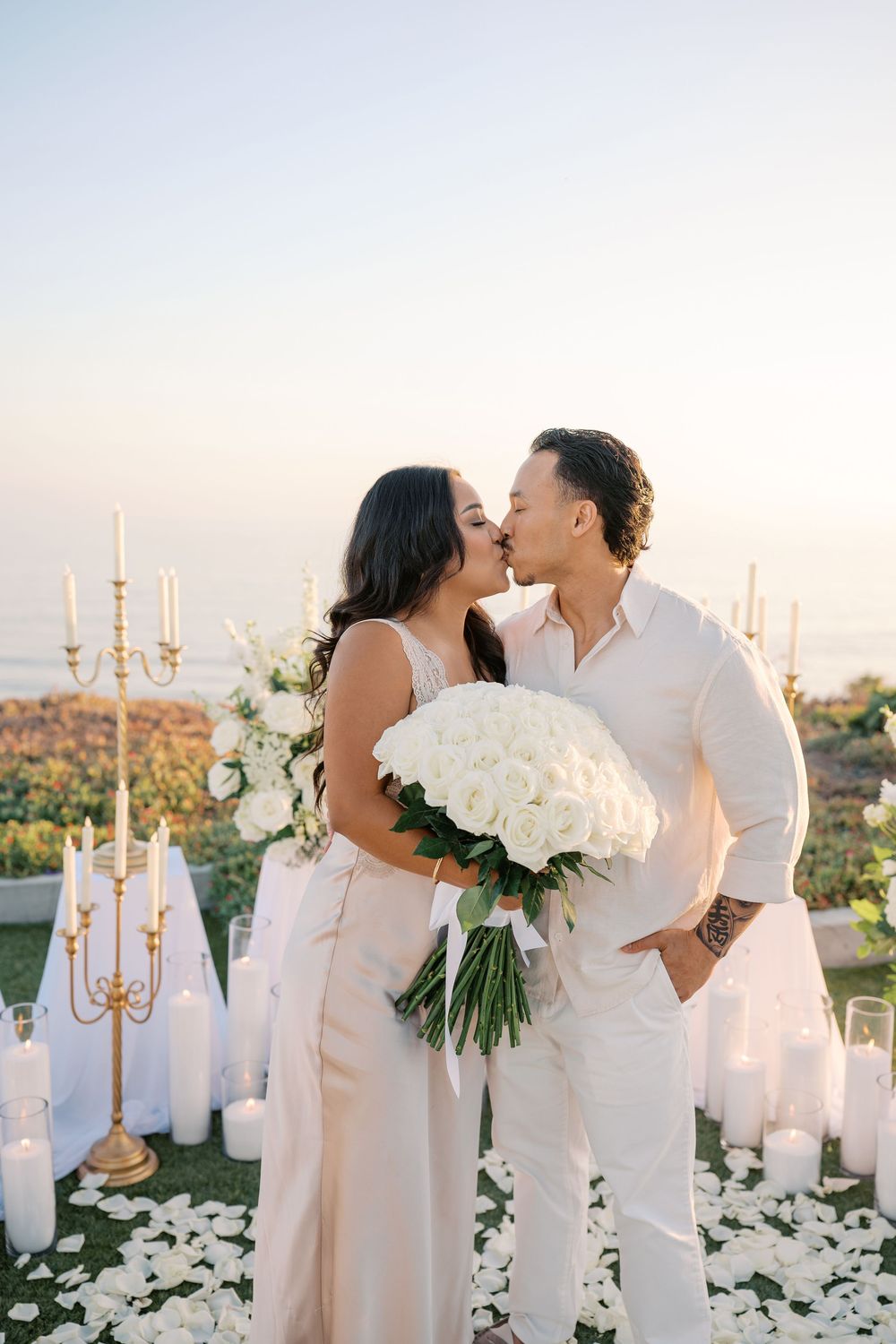 Couple kissing during romantic outdoor proposal with white roses, candles, and ocean sunset backdrop.