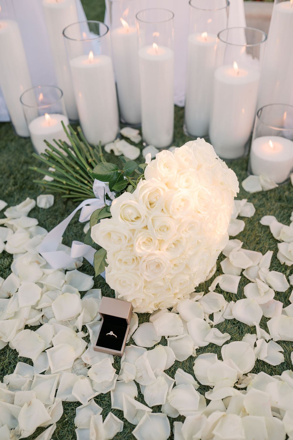 White rose bouquet with ring box, rose petals, and lit candles on grass for a romantic wedding proposal setup.