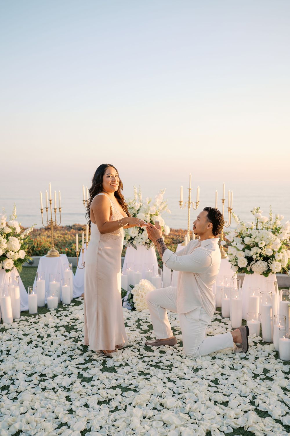 Man proposes on one knee amid white roses, candles, and floral arrangements at a romantic oceanside sunset setting.