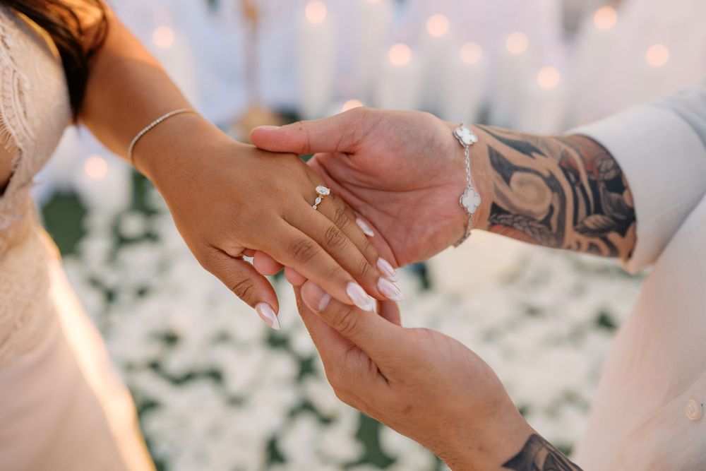 Groom with tattoos placing a diamond engagement ring on brides finger during a romantic wedding ceremony.