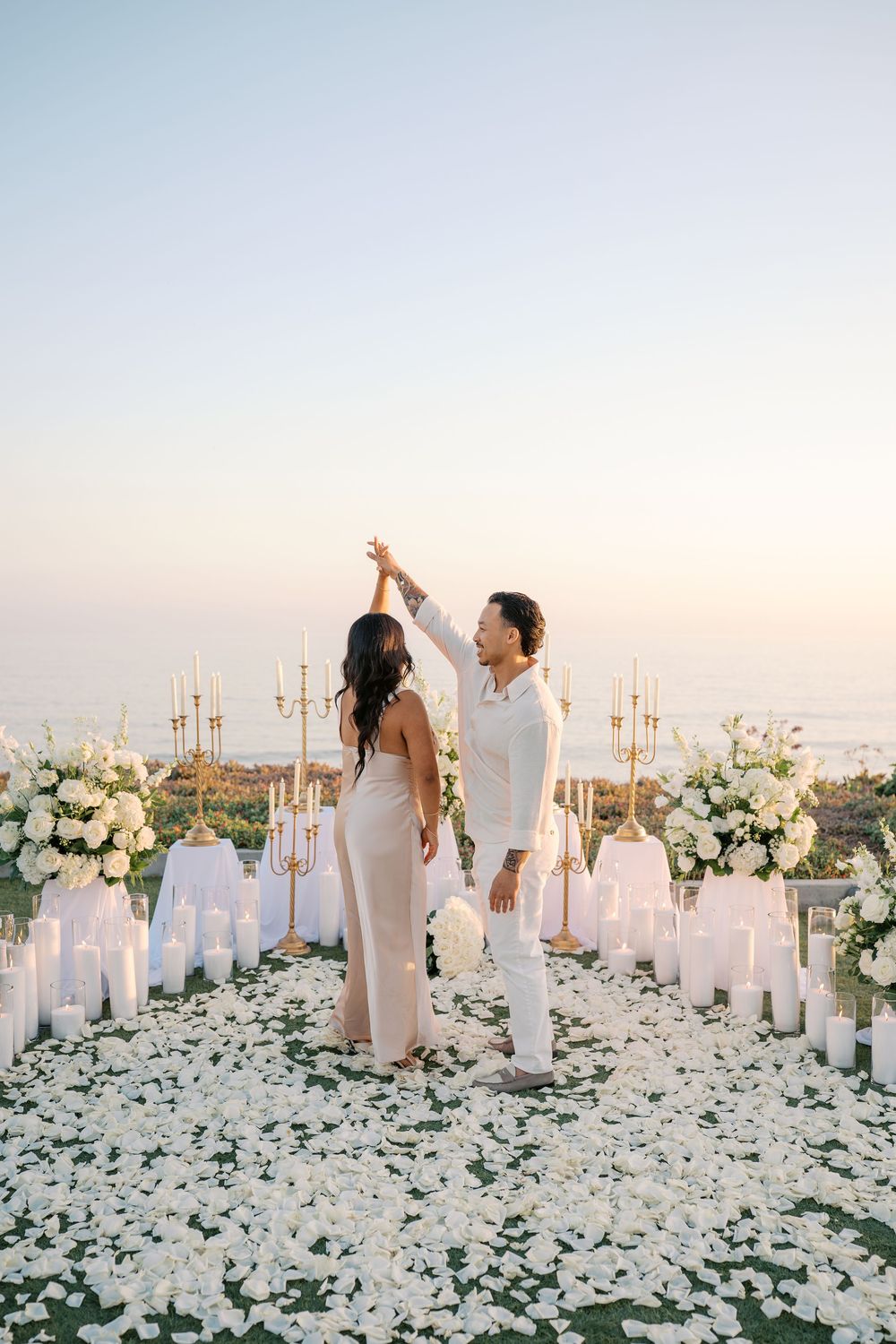 Couple dancing at romantic outdoor proposal setup with white flowers, candles, and ocean view at sunset.