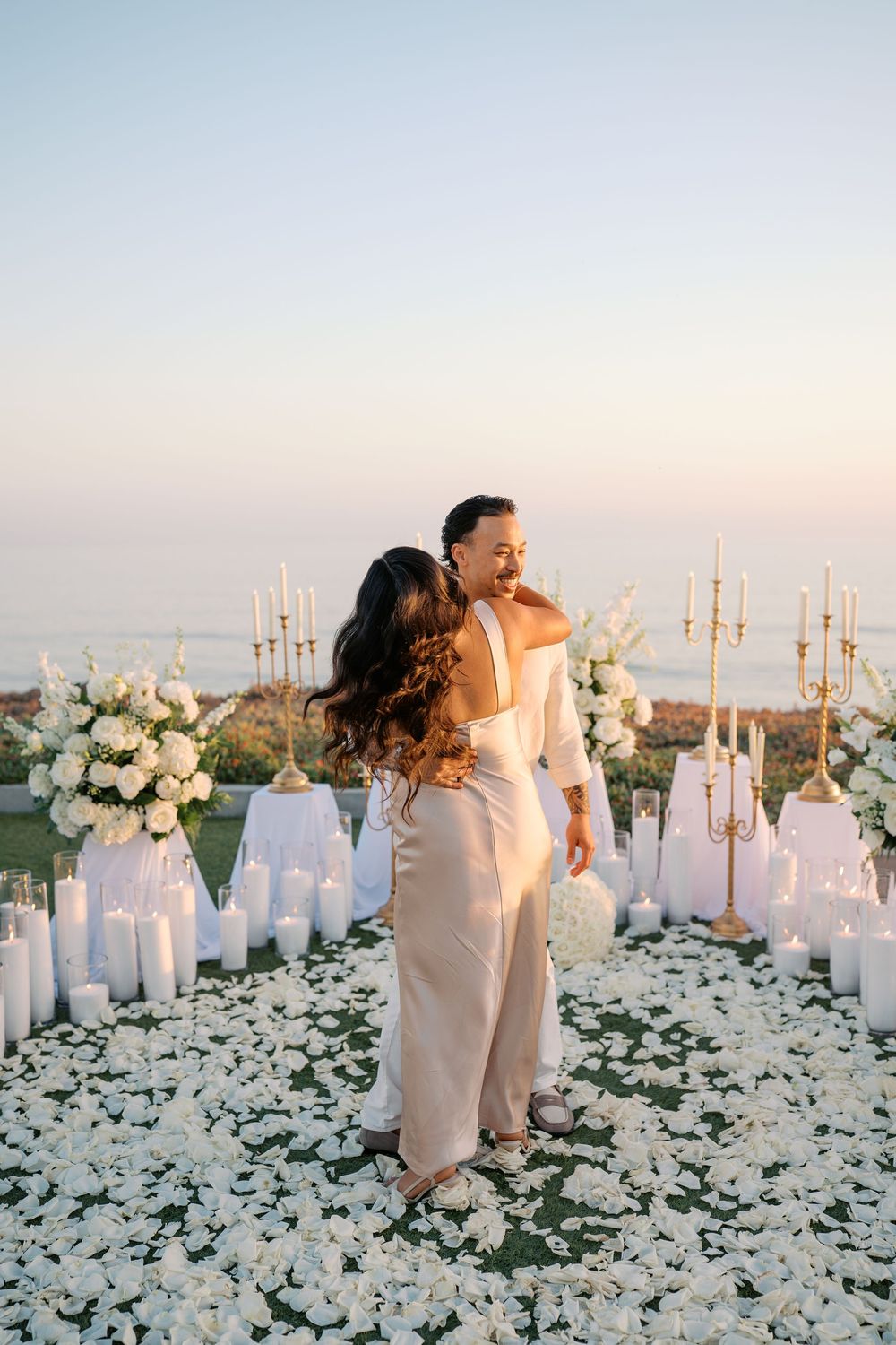 Couple embracing at romantic sunset proposal setup with white flowers, candles, and rose petals by the ocean.