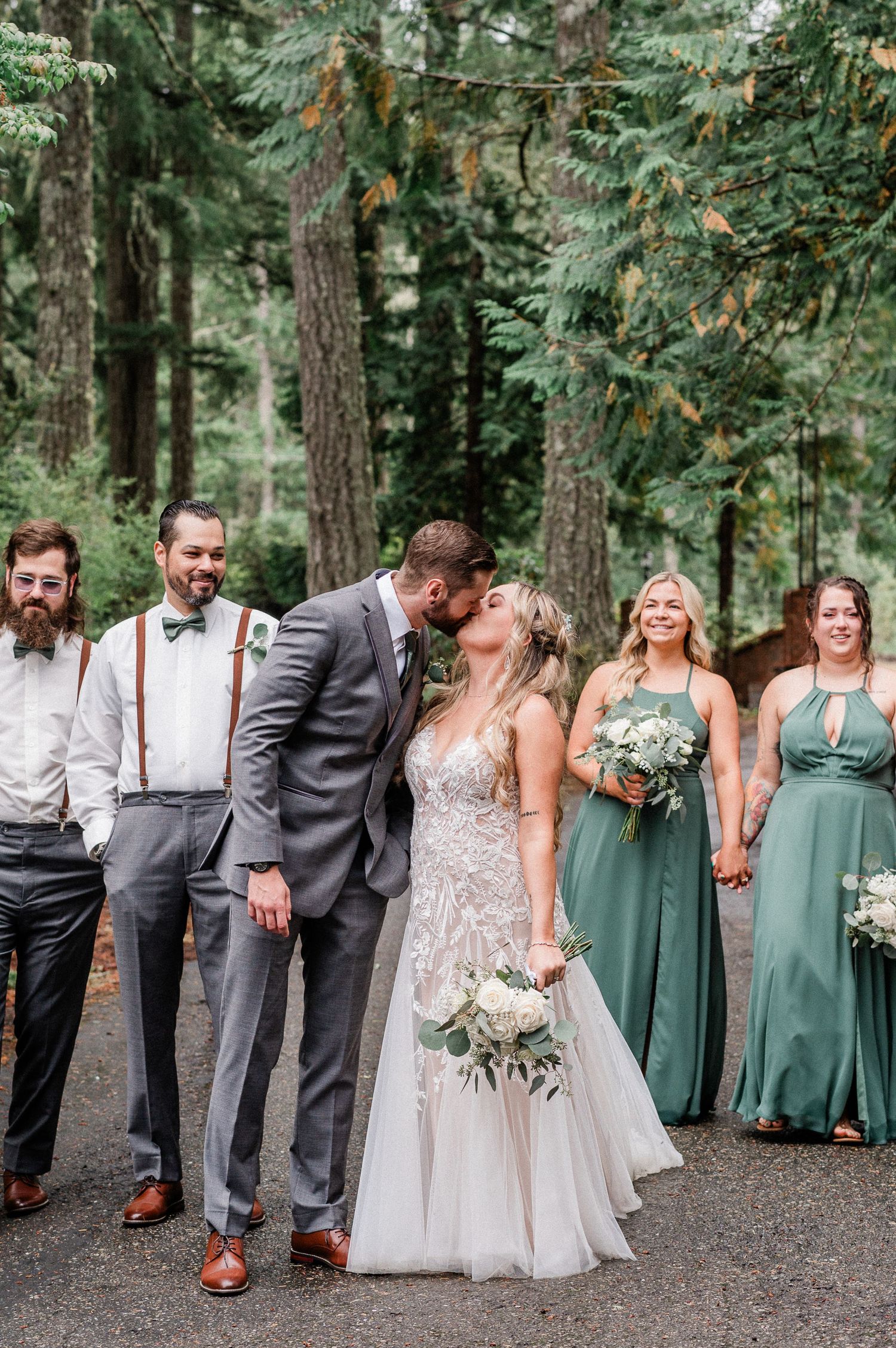 Bride and groom share a kiss surrounded by wedding party in green dresses and gray suits in a forest setting.