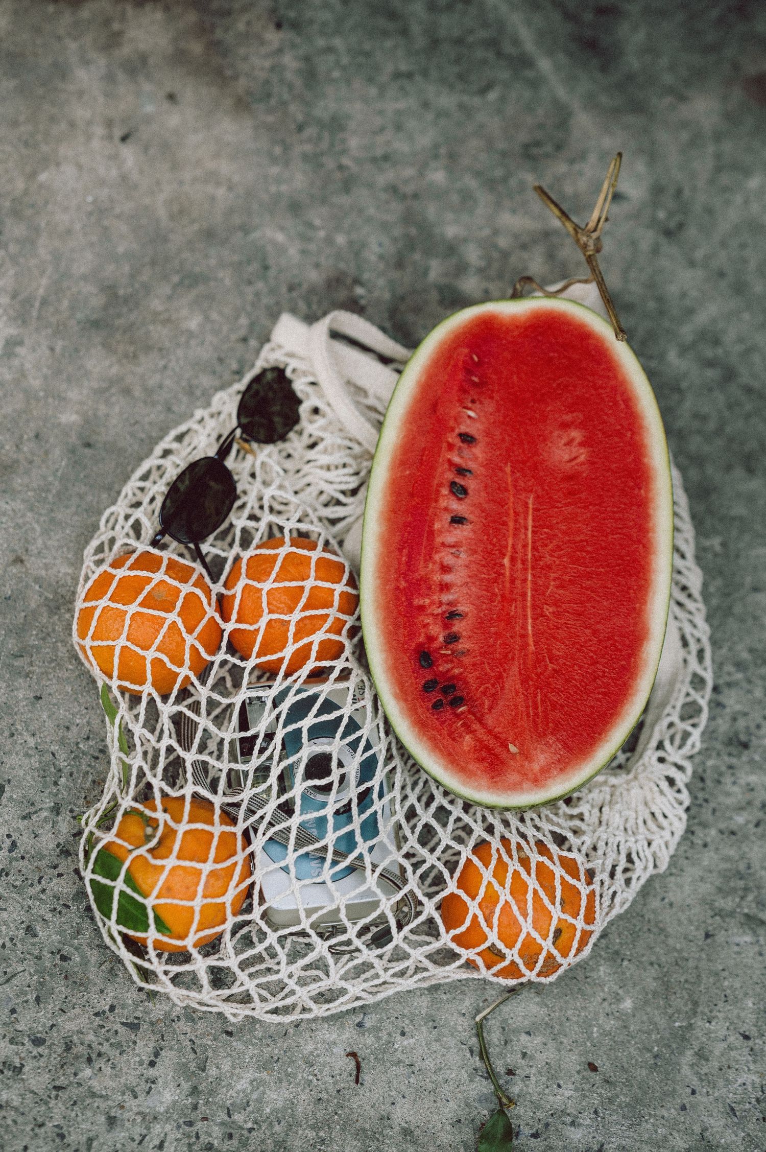 Flat lay of a mesh bag with watermelon and oranges on concrete at a Vietnamese street market — editorial prop styling