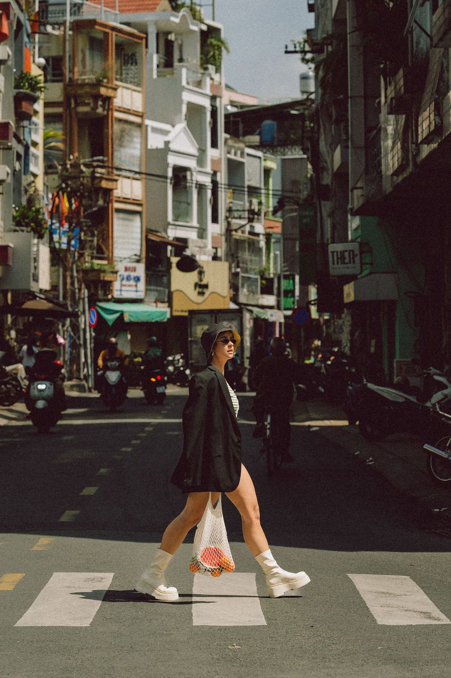 Full-length editorial portrait of a woman crossing a street in Ho Chi Minh City wearing a black blazer and white boots — urban fashion photo