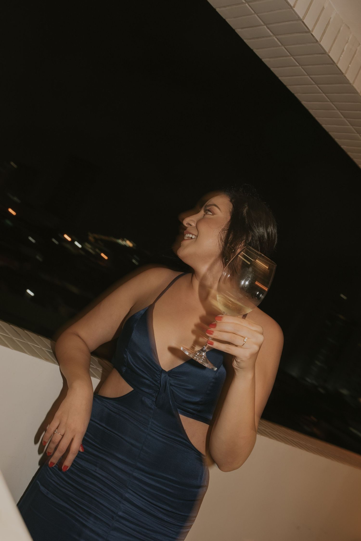 Woman in navy blue dress laughs while holding a drink at a nighttime celebration.