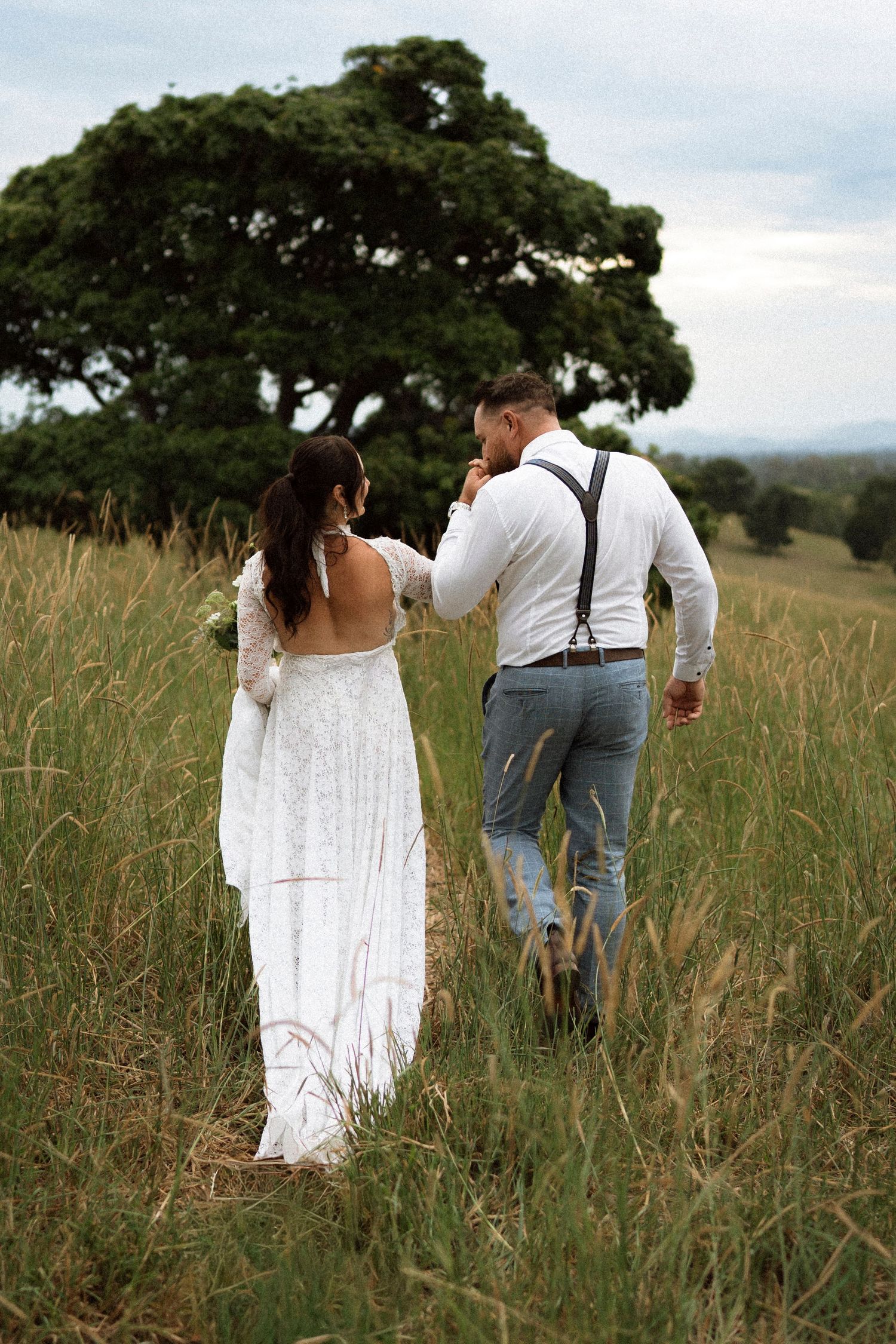 Bride and groom walking through tall grass field toward large tree, bride in backless lace gown, groom in suspenders.