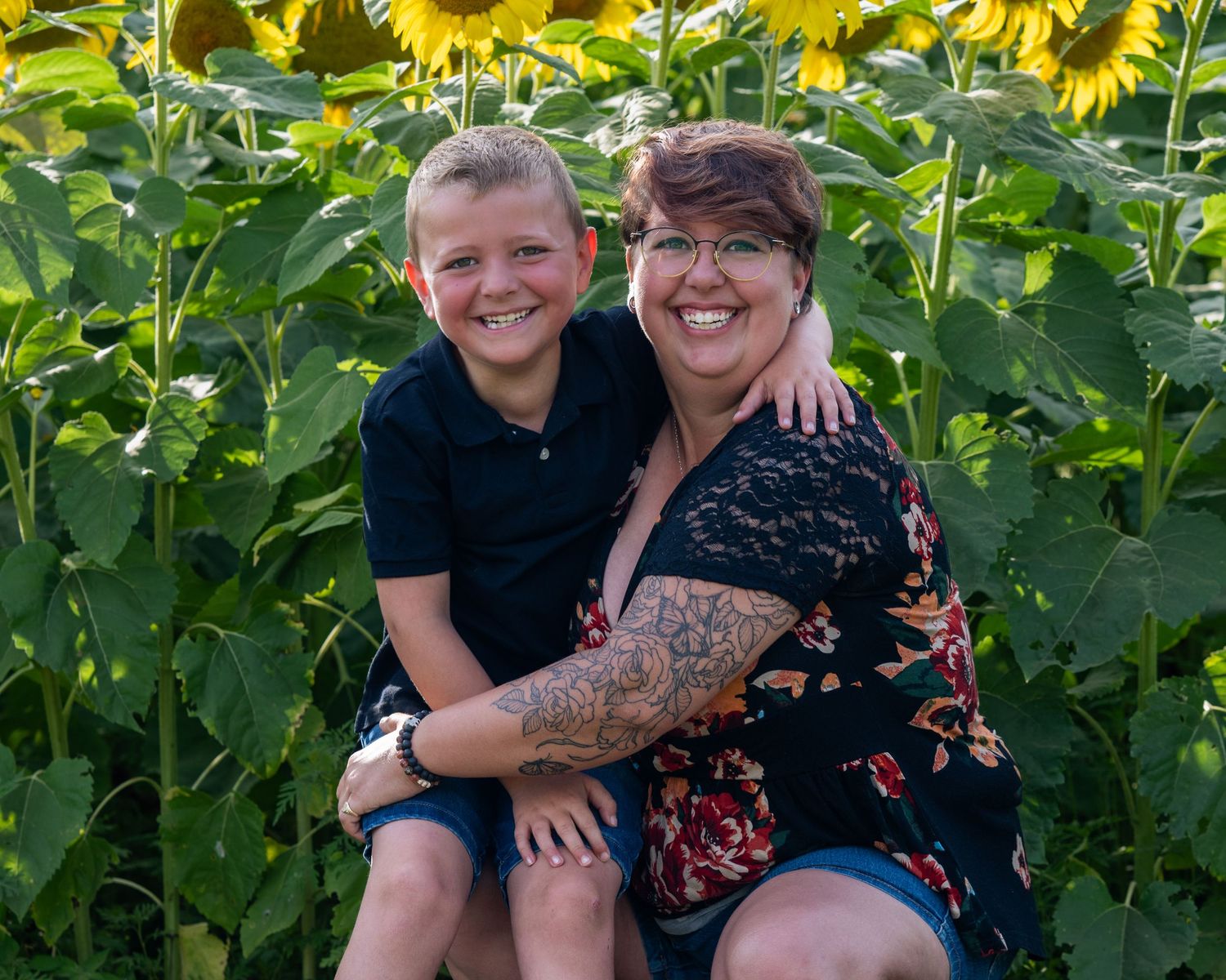 Smiling mother and son embracing outdoors in front of tall sunflowers on a bright summer day.