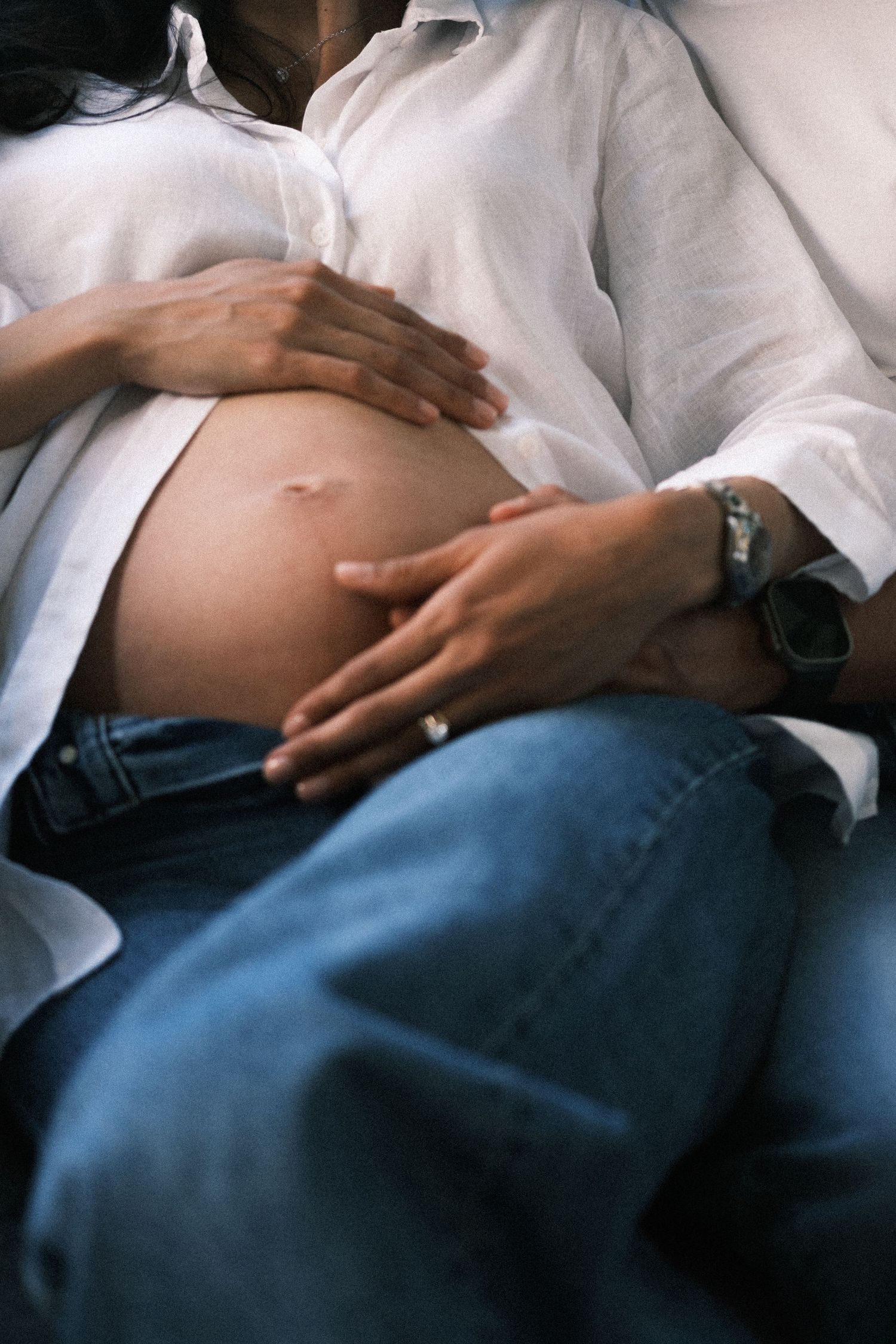 Pregnant woman in white shirt and jeans with partner's hands gently cradling her bare baby bump.