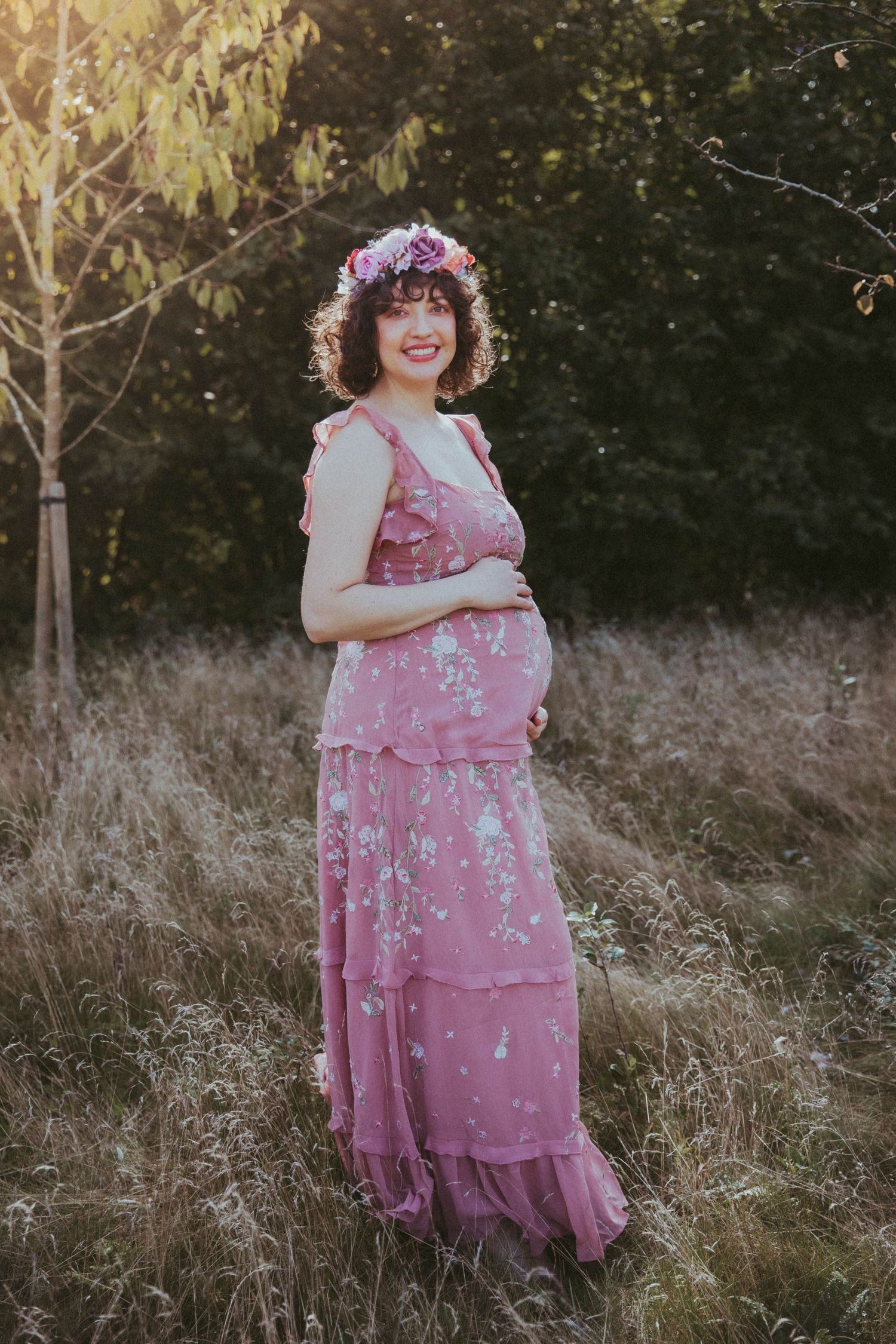 Smiling pregnant woman in pink floral maxi dress and flower crown standing in a sunlit field. During a location maternity session in Bexley