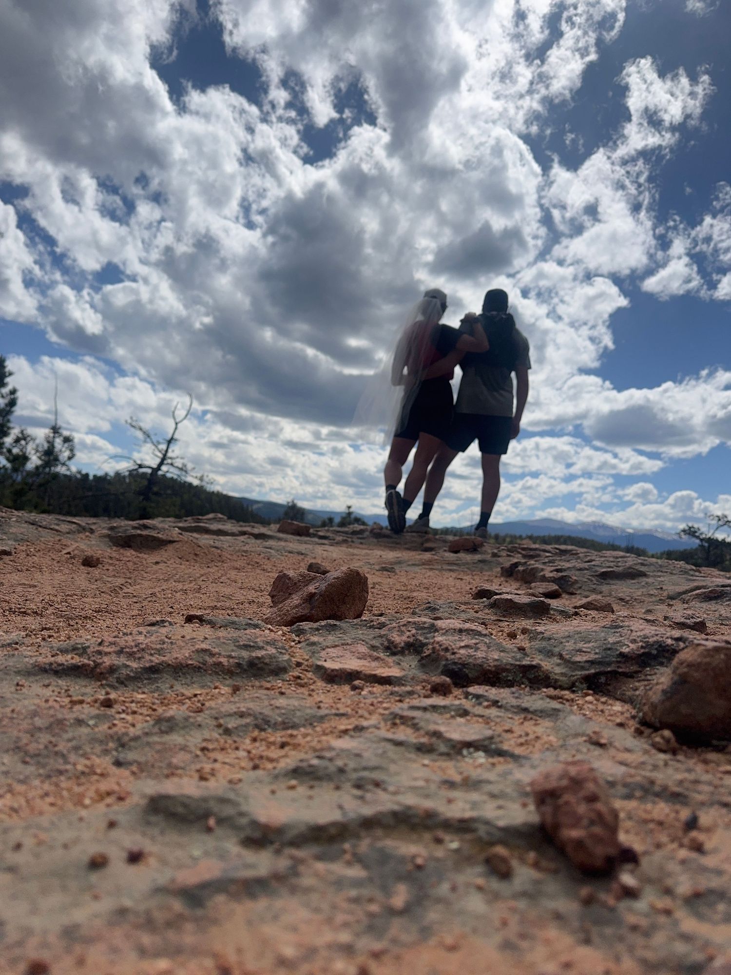 Two hikers stand arm in arm on a rocky desert trail under a dramatic cloudy sky with mountain views in the distance.