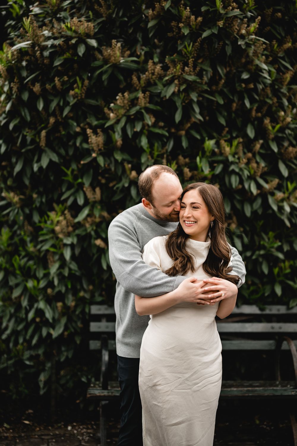 A couple embraces warmly in front of a lush green hedge, the woman in a white dress smiling joyfully.