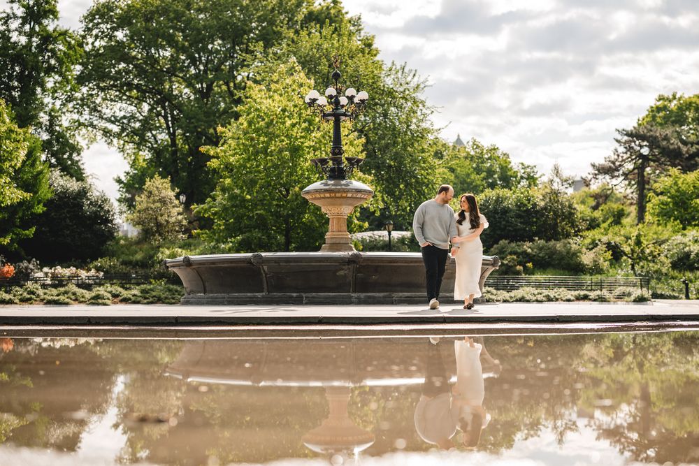 Couple embracing near a stone fountain in a lush green park, reflected in still water below.