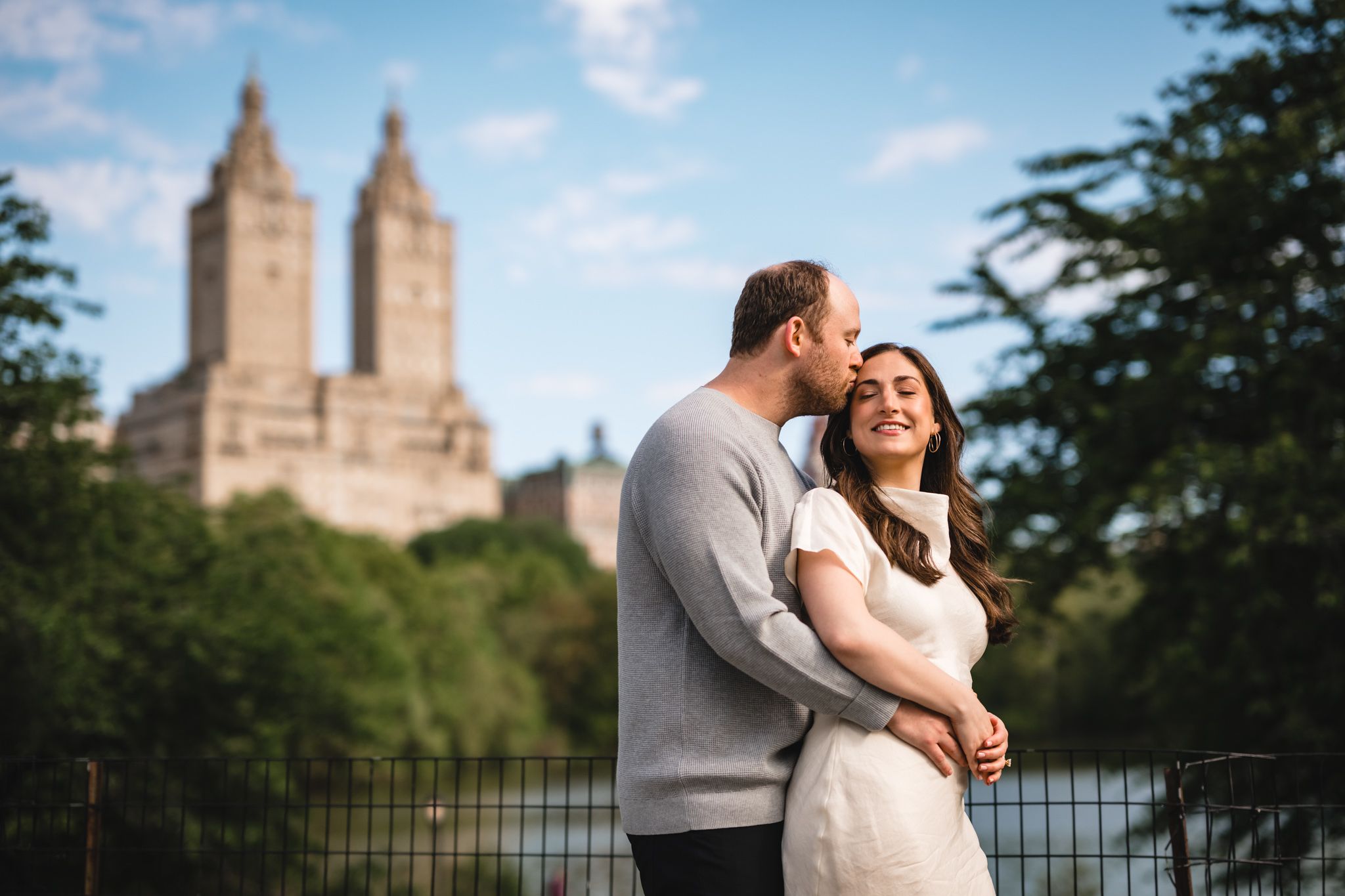 Couple embracing in Central Park with iconic San Remo building towers visible against a blue sky.