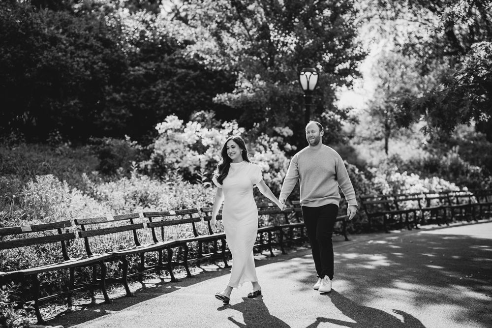 A couple walks hand-in-hand along a sunlit park path, surrounded by lush greenery, in a black-and-white photo.