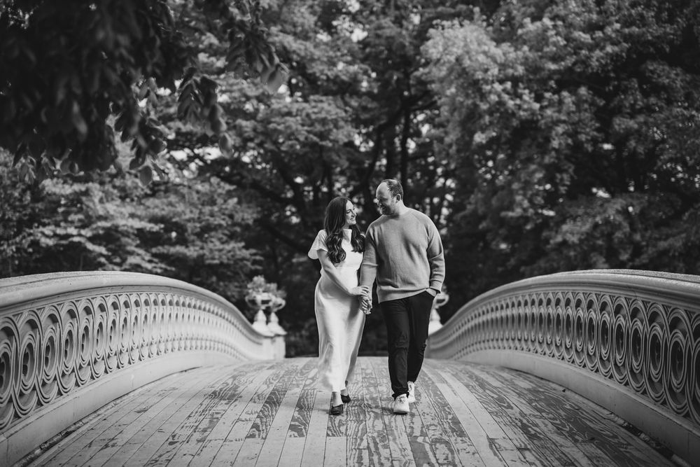 Couple walking hand in hand on Bow Bridge in Central Park in a romantic black and white photo.