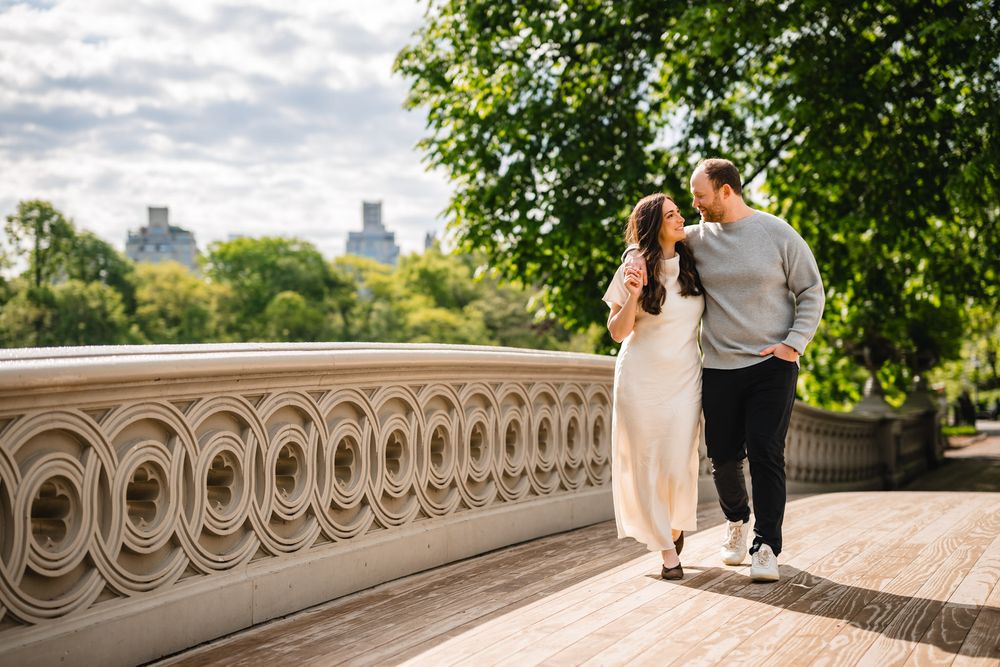 Couple walking on Bow Bridge in Central Park, NYC, surrounded by lush green trees on a cloudy day.
