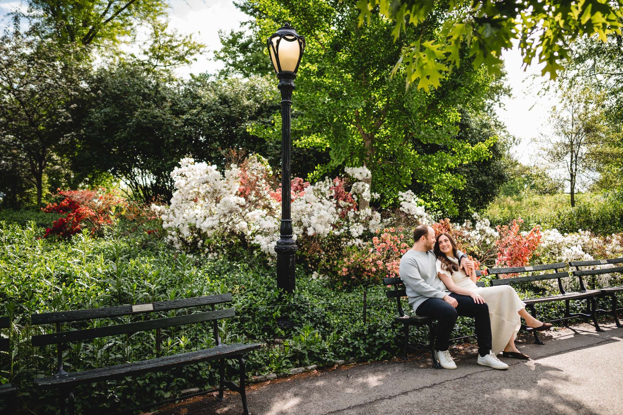 Couple sitting together on a park bench surrounded by colorful blooming flowers and green trees.