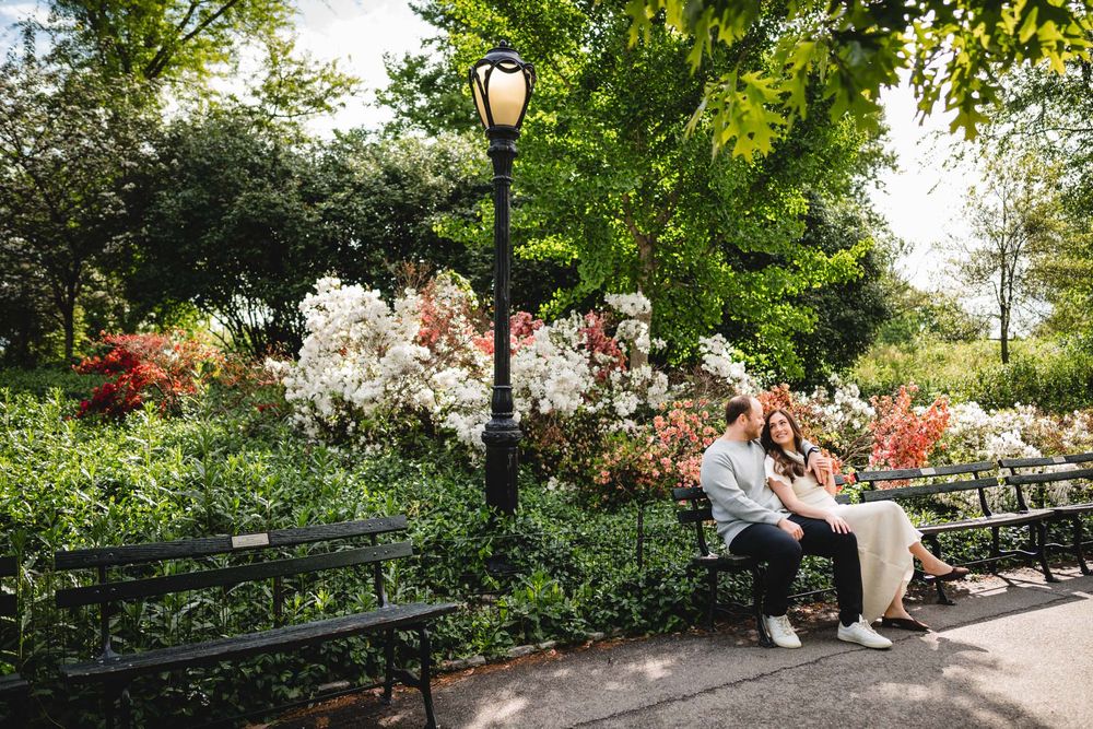 Couple sitting together on a park bench surrounded by colorful blooming flowers and green trees.