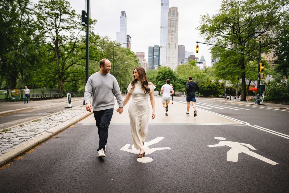 Couple holding hands crossing a street in Central Park, NYC, with skyscrapers and lush trees in the background.