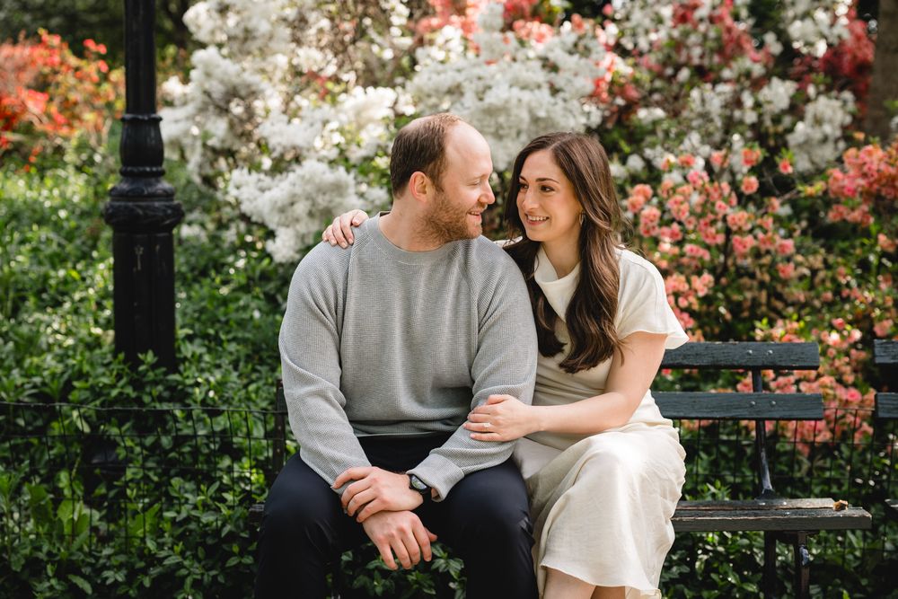 Couple sitting on a park bench surrounded by blooming spring flowers, sharing a romantic moment outdoors.