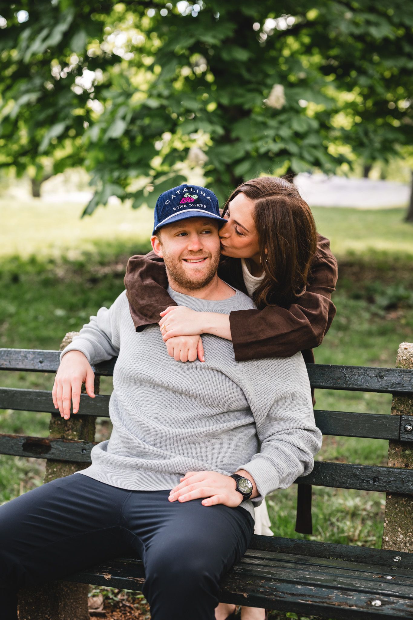 Couple sharing a tender moment on a park bench, woman kissing man's cheek while embracing him from behind.