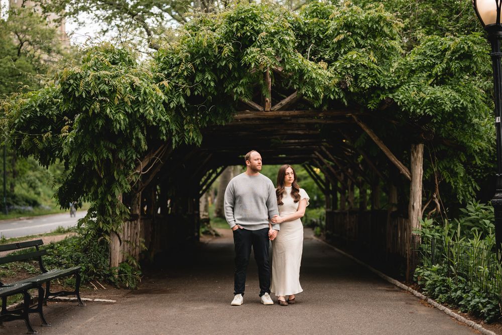 Couple holding hands under a lush green pergola tunnel in a park, surrounded by trees and benches.
