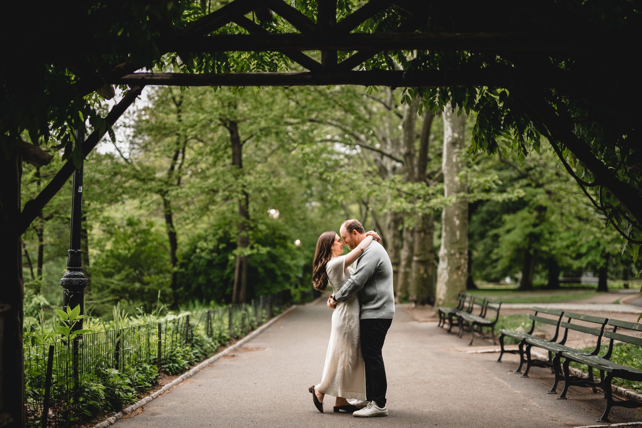 Couple sharing a romantic kiss under a vine-covered pergola in a lush green park with benches lining the path.