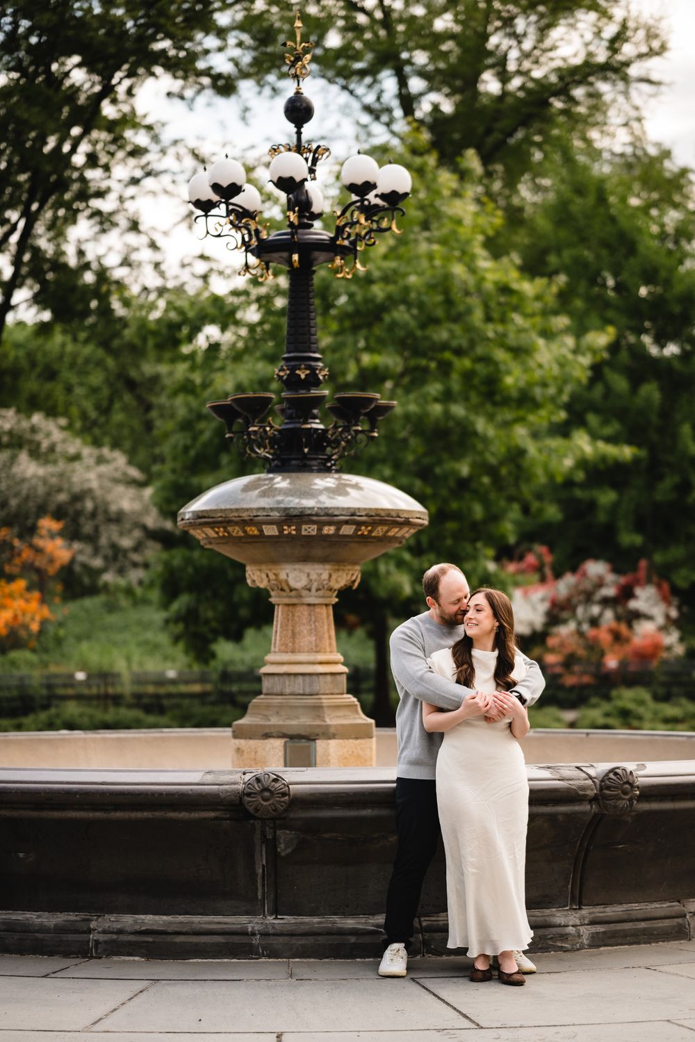 Couple embracing in front of an ornate stone fountain surrounded by lush greenery in a park.