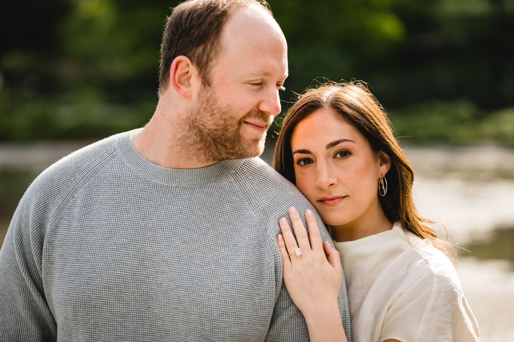 A couple poses closely together outdoors, woman resting head on man's shoulder, both smiling softly.