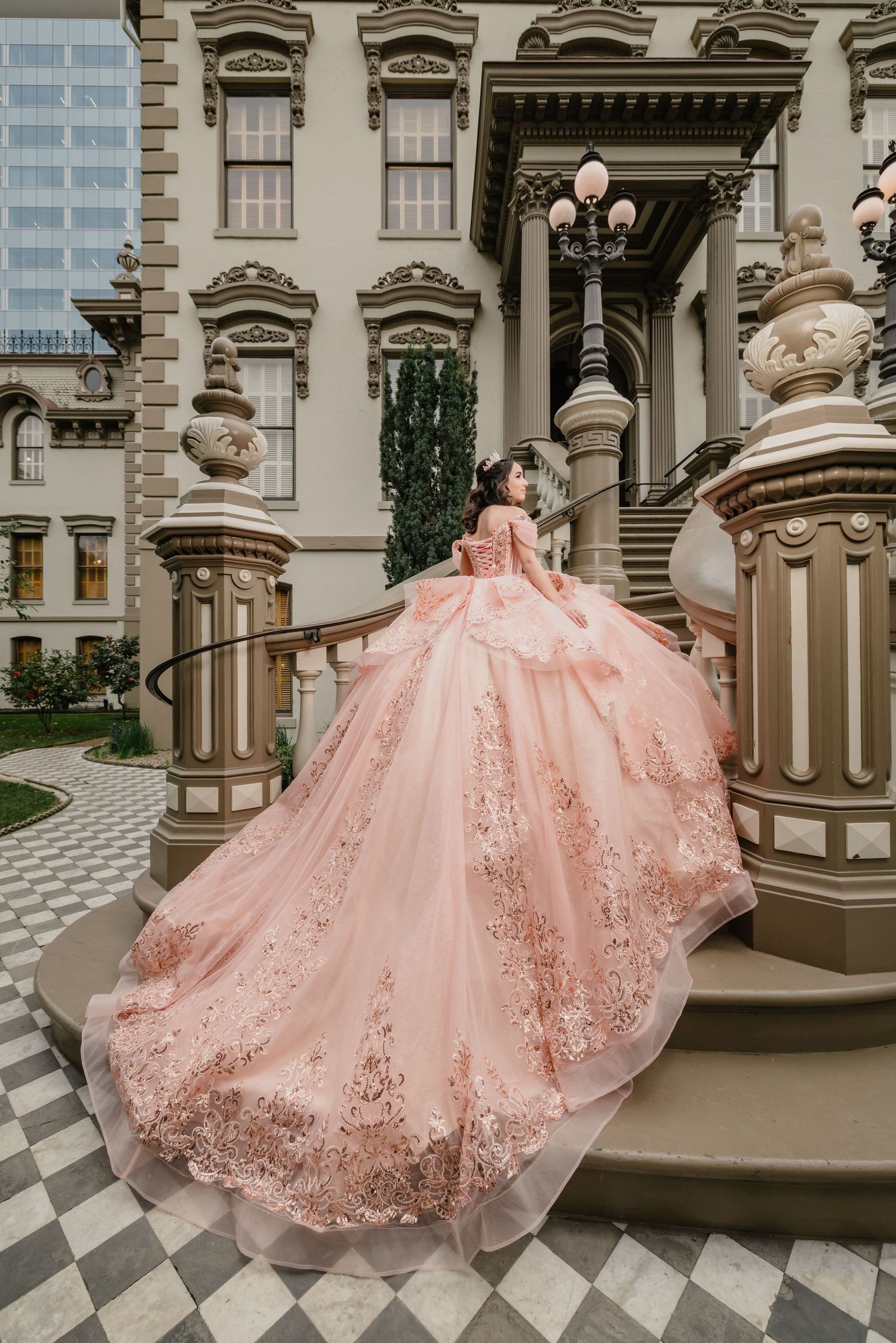 Woman in blue ball gown poses by a pond in front of a white castle-like mansion on a sunny day.
