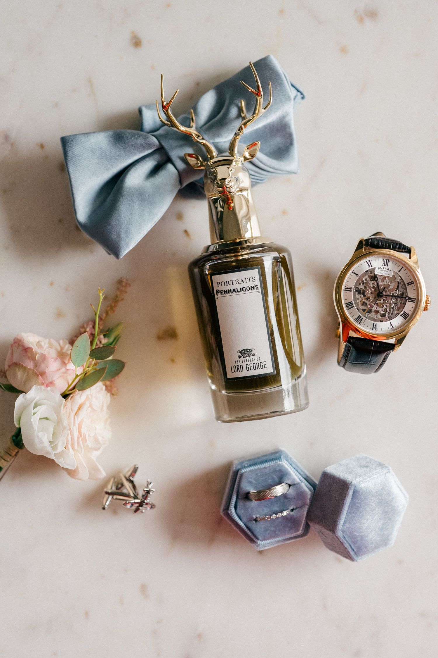 Elegant wedding flat lay with Penhaligon's perfume, gold watch, cufflinks, ring box, blue bow, and floral bouquet on marble.
