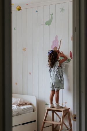 Girl standing on a wooden step stool decorating a white paneled bedroom wall with colorful mermaid stickers.