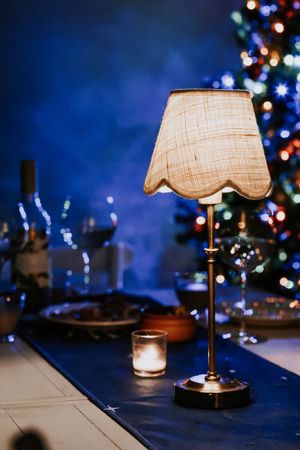 Elegant table lamp with burlap shade on holiday dinner table with Christmas tree bokeh lights in background.