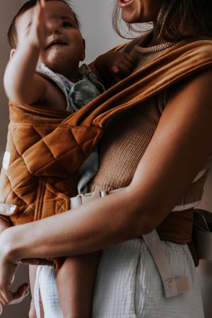 A mother carrying her happy baby in a rust-colored quilted baby carrier, both wearing neutral tones.