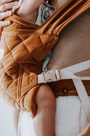 Close-up of a brown quilted baby carrier with buckle strap worn by a parent holding an infant.