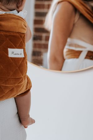 Baby in brown quilted Mumma carrier, mother reflected in round mirror against white wall.