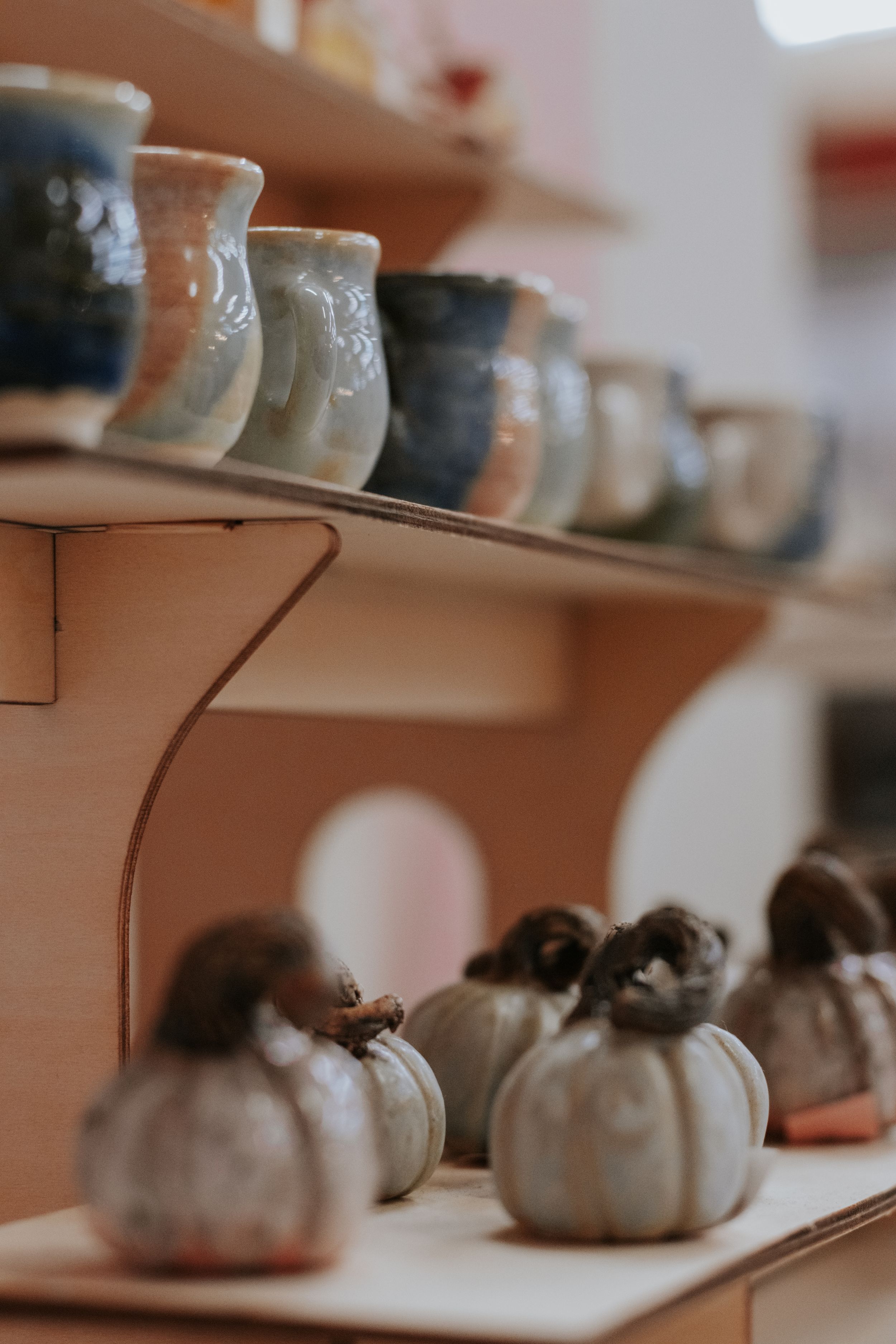 Handmade ceramic mugs on wooden shelves with glazed pottery pumpkin figurines displayed in a cozy artisan shop.
