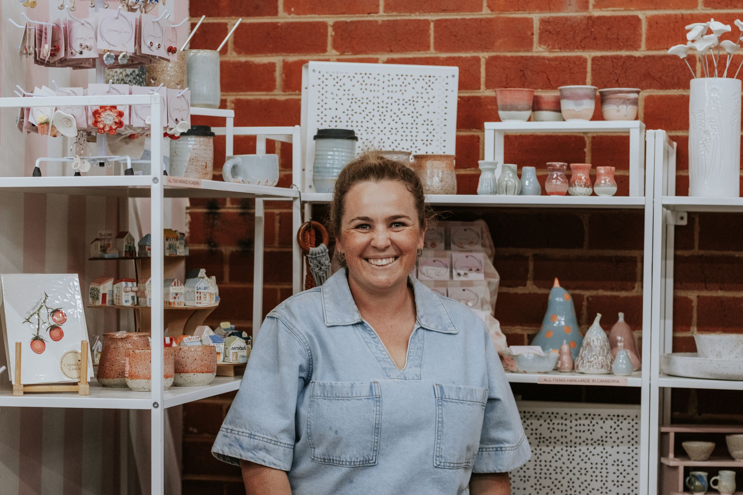 Smiling woman in blue denim shirt standing in front of shelves displaying handmade ceramic pottery in a cozy shop.