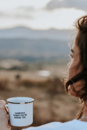Woman holding a white enamel mug reading Canberra Thinks You're Boring Too with scenic mountain backdrop.