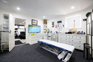 Modern physiotherapy treatment room with massage table, white dresser, TV screen, and medical equipment on dark carpet.