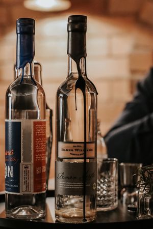 Two clear spirit bottles from Baker Williams Distillery displayed on a dark table with glassware in soft bokeh lighting.
